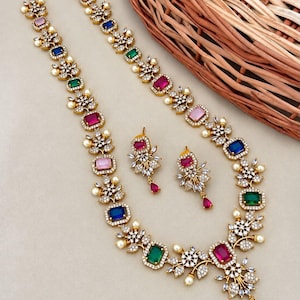 May include: A gold-toned necklace and matching earring set featuring rectangular multi-colored gemstones in pink, blue, and green. The jewelry is accented with small clear crystals and tiny white pearls arranged in floral-inspired metal settings.