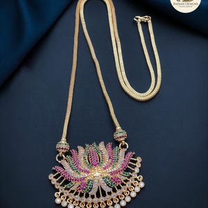 May include: Gold-toned necklace featuring a pendant shaped like a lotus flower. The pendant is adorned with pink, green, and white gemstones. The necklace has a delicate chain and a clasp closure. The jewelry is displayed on a dark blue surface.