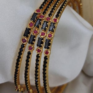 May include: A set of four gold-tone bangles featuring alternating black rectangular stones and small round red stones. The metal bands have a polished finish, and the stones are set in a repeating pattern along the outer circumference of each bracelet.