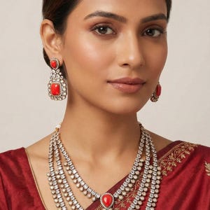 May include: A three-strand necklace, earrings, and headpiece set. The jewelry features red gemstones and clear stones set in a gold-colored metal. The necklace has a large, rectangular red stone pendant. The earrings are square-shaped with red stones. The headpiece has a teardrop-shaped red stone.