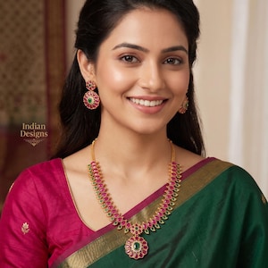May include: A gold-toned necklace and earring set featuring ruby-colored and green gemstones. The necklace has a floral pendant and matching earrings. The image also includes the text "Indian Designs".