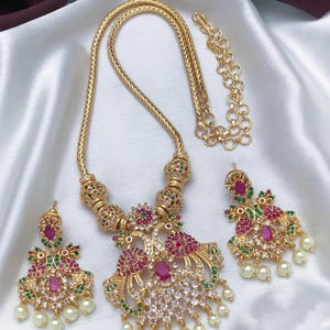 May include: A gold-tone necklace and earring set featuring peacock motifs adorned with pink, green, and clear stones. The necklace has a thick chain with decorative beads and a large pendant, while the matching earrings include dangling white pearls.