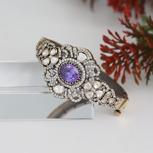 May include: A gold-toned bangle bracelet with a purple gemstone surrounded by clear stones. The bracelet has a delicate, intricate design.