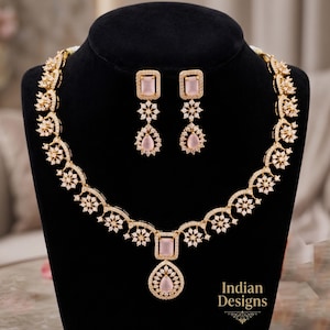 May include: A gold-toned necklace and earring set with pink gemstones and clear crystals. The necklace features a floral design with a square pendant and a teardrop-shaped gemstone. The earrings have a matching design. The set is displayed on a black bust.