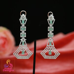 May include: A pair of silver dangle earrings with a geometric design. The earrings feature clear cubic zirconia stones and green accents.