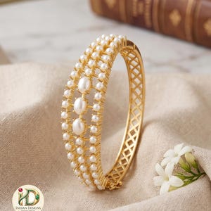 May include: A gold-toned bangle bracelet adorned with multiple rows of small pearls and three larger pearls. The bracelet has a lattice design on the sides and is displayed on a beige fabric. The jewelry is from Indian Designs.