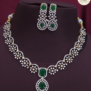 May include: A gold-coloured necklace and earring set with emerald green gemstones and clear crystals. The necklace features a floral design and a large emerald pendant. The earrings have a matching design with emerald-cut stones.