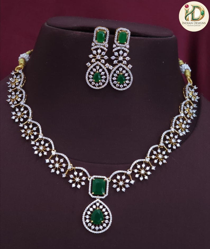 May include: A gold-coloured necklace and earring set with emerald green gemstones and clear crystals. The necklace features a floral design and a large emerald pendant. The earrings have a matching design with emerald-cut stones.