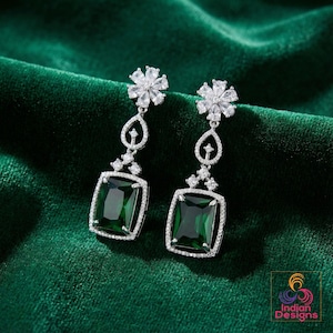 May include: A pair of silver-toned drop earrings with a floral design at the top and a rectangular, emerald-green gemstone at the bottom. The earrings are set against a dark green velvet background.