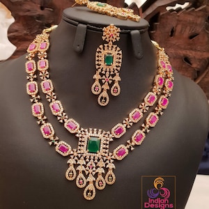 May include: A two-tiered gold necklace with pink and green stones. The necklace has a pendant with a green stone and a teardrop design. The necklace is displayed on a black mannequin. The image also includes a pair of matching earrings.