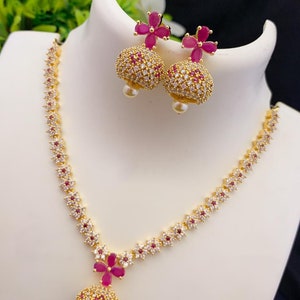 May include: A gold necklace with a pendant and matching earrings. The necklace and earrings are adorned with small white stones and pink flowers.