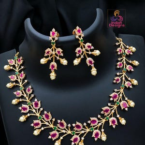 May include: A gold-toned necklace and earring set. The jewelry features a floral design with ruby-colored stones, small green accents, and white pearl-shaped drops. The set is displayed on a black mannequin head.