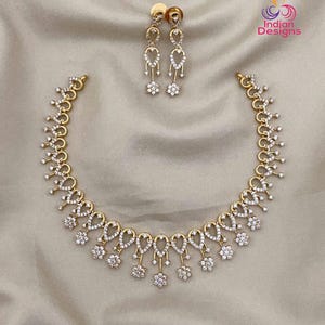 May include: A gold-toned necklace and matching pair of drop earrings, each featuring a series of teardrop-shaped links encrusted with small clear stones and accented by dangling floral-shaped charms set with clear stones, Indian Designs logo.