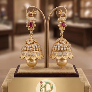 May include: Gold-toned, ornate earrings with a peacock design. Each earring features a ruby accent, clear stones, and a large, golden pearl. The earrings are displayed on a stand with the "Indian Designs Fashion Jewelry" logo.