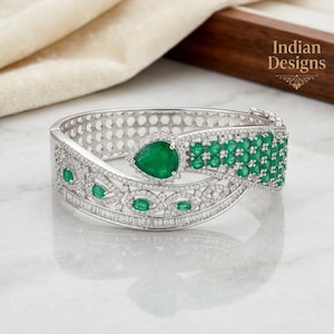 May include: A silver-toned hinged bangle bracelet featuring a large pear-shaped green gemstone, multiple smaller round and marquise-cut green stones, and numerous tiny clear pavé-set stones. The bracelet displays the text Indian Designs.