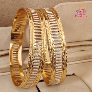 May include: Two gold bangles with a white and gold striped design. The bangles have a textured surface and are displayed on a neutral background. The bangles are a piece of jewelry.