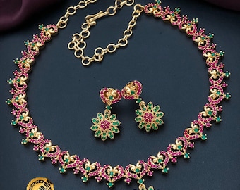 May include: A gold-toned necklace and earring set featuring intricate heart-shaped links adorned with small pink and green stones. The necklace includes a central flower-shaped pendant, and the earrings are matching flower designs. Text reads Indian Designs and Premium Quality Micro Plated AD Necklace.