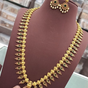 May include: A gold-colored necklace and matching earrings set. The necklace features a series of ornate, leaf-shaped pendants with green and red accents. The earrings have a similar design, with a central green stone and small white pearls.