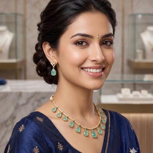 May include: A gold necklace featuring multiple teardrop-shaped light green stones and small clear accent stones, paired with matching teardrop-shaped light green stone earrings. The jewelry set is worn with a dark blue garment with gold embroidery.
