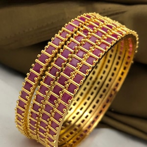 May include: Set of four gold bangles with pink square-shaped stones. The bangles are stacked on top of each other.