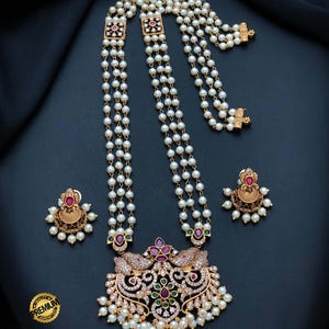 May include: A multi-strand pearl necklace featuring a large gold-tone pendant with embedded red and green stones, clear crystals, and small dangling pearls. The set includes matching gold-tone stud earrings with red stones and pearl accents.