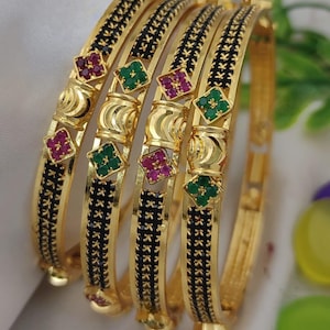 May include: Four gold bangles with black and gold detailing. Each bangle features a repeating black pattern and is adorned with square-shaped accents of red and green gemstones. The bangles are displayed on a white surface.
