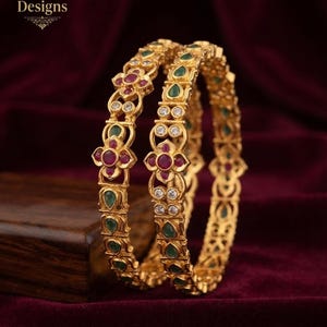 May include: Two gold bangles featuring Indian Designs text, adorned with circular red stones, teardrop-shaped green stones, and small clear white stones arranged in a repeating floral and geometric pattern along the metallic band surface.