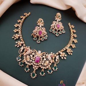 May include: Gold-toned necklace and earring set with intricate peacock and floral designs. The necklace features a central ruby-colored stone and is adorned with clear and emerald-green accents. The earrings mirror the necklace's design, creating a cohesive jewelry set.