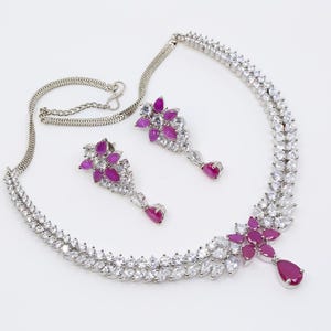 May include: A silver necklace and earrings set with a double row of clear cubic zirconia stones and pink accents. The necklace has a floral design with a teardrop pendant.