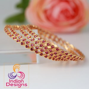 May include: Three gold-toned bangles featuring a continuous wave-like metal structure set with small, round, deep pink stones. The bangles are arranged in a stack, and the image includes a logo with the text Indian Designs in the bottom corner.