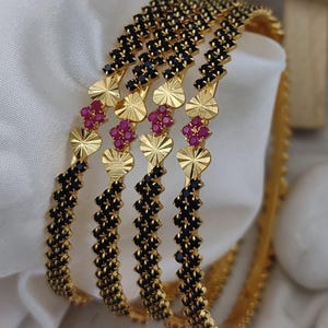 May include: A set of four gold bangles featuring a repeating pattern of black stones and small, textured gold heart-shaped accents. Each bangle is also decorated with a cluster of four small, vibrant pink stones arranged in a square formation.