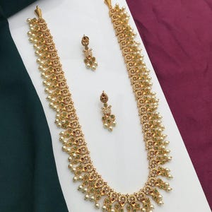 May include: A gold-toned necklace and matching earrings set. The necklace features a detailed design with small red and green accents, and is adorned with faux pearls. The earrings have a similar design. The jewelry set is displayed on a white surface.