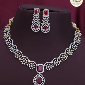 May include: A gold-coloured necklace and earring set. The necklace features a large, rectangular ruby-coloured stone pendant with a teardrop-shaped stone below. The earrings and necklace are adorned with small, clear crystals.