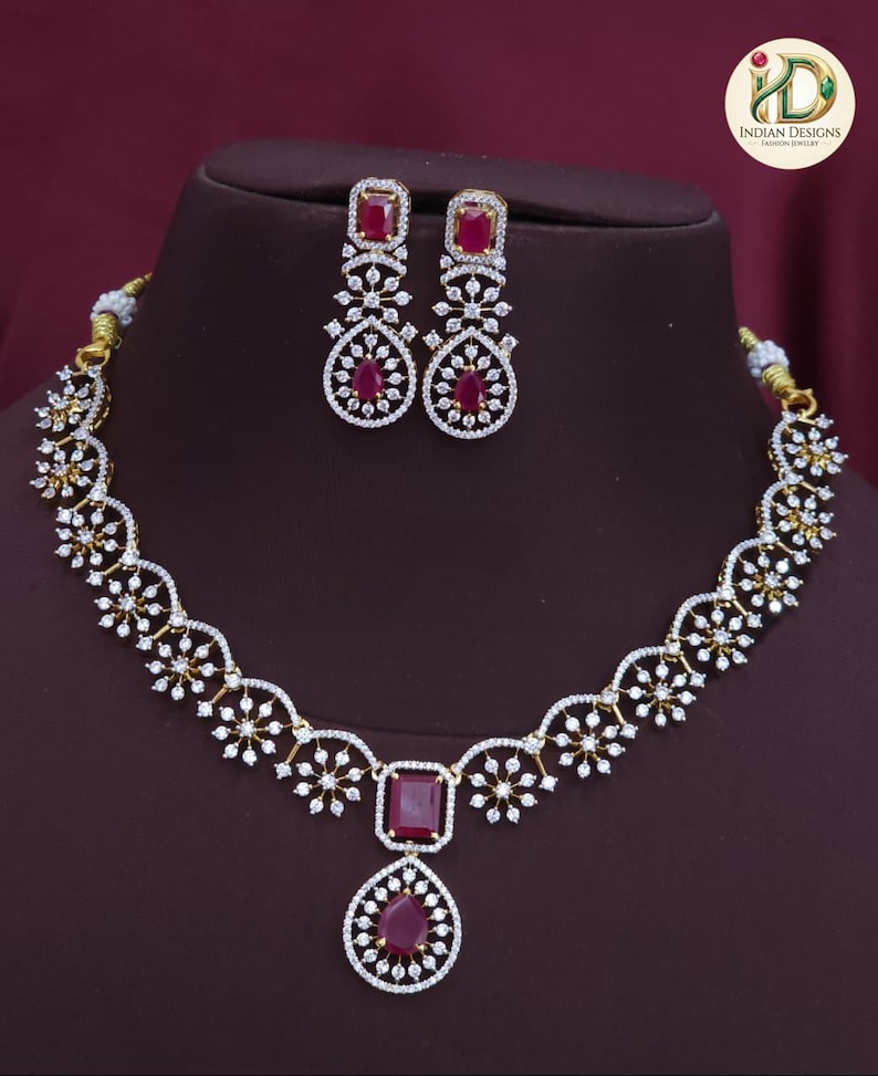May include: A gold-coloured necklace and earring set. The necklace features a large, rectangular ruby-coloured stone pendant with a teardrop-shaped stone below. The earrings and necklace are adorned with small, clear crystals.