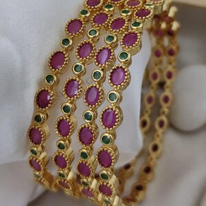 May include: A set of three gold-toned bangles featuring a repeating pattern of oval-shaped magenta stones and small round green stones. Each stone is set within a textured, beaded gold metal frame, creating a continuous decorative band design.