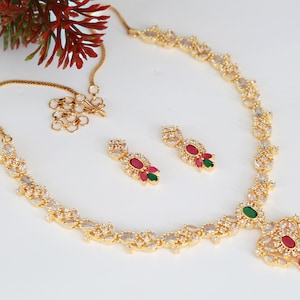 May include: A gold-toned jewelry set featuring a necklace and matching earrings. The necklace and earrings are adorned with clear and red gemstones, with a green accent stone on the necklace. The set is displayed on a white surface.