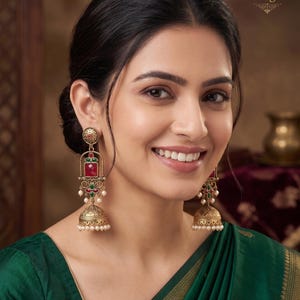 May include: Gold-toned jhumka earrings featuring a circular stud, a rectangular red stone, and a bell-shaped drop. The design includes small green accents and dangling white beads. The text Indian Designs is visible in the upper right corner.