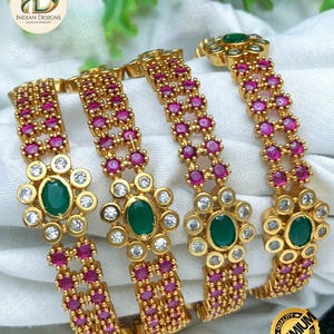 May include: Four gold-toned bangles featuring a central floral motif with an oval green stone surrounded by small white stones. The bands are decorated with two parallel rows of small round pink stones. The logo ID Indian Designs is visible.