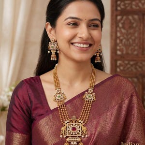 May include: A woman wearing a gold necklace and matching earrings. The necklace has multiple strands and features square and rectangular pendants with red and green stones. The earrings have a similar design. The woman is wearing a maroon top.