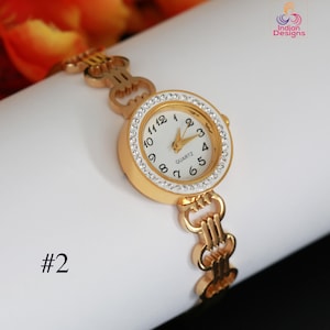May include: A gold-toned quartz watch with a white face and crystal-studded bezel. The watch features black numerals and gold hands. The watch band is also gold-toned with a decorative link design. The number "#2" is printed on the lower left.