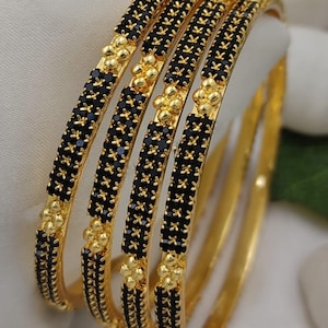 May include: A set of four gold-tone bangles featuring a central band of small black stones. Each bangle is accented with clusters of polished gold-tone beads at regular intervals, creating a textured, repeating pattern along the circular frame.