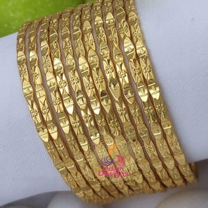 May include: A stack of gold-colored bangles with an intricate, repeating pattern. The bangles are arranged on a white surface, with a few green apples visible in the background. The bangles have a shiny, reflective surface.