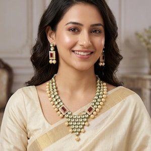 May include: A multi-layered necklace and matching rectangular drop earrings featuring clear stones, deep red rectangular accents, and cream-colored pearls. The jewelry set is crafted with a gold-toned metal finish and a symmetrical, tiered design.