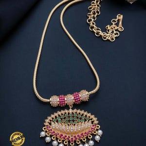 May include: A gold-toned necklace featuring a snake chain with three cylindrical beads encrusted with pink and white stones. The large oval pendant displays a lotus-inspired design with green, pink, and white stones, finished with hanging white pearls.