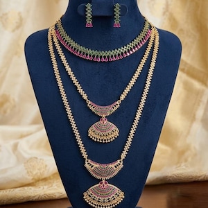 May include: A gold-toned jewelry set featuring a choker necklace, two layered necklaces with fan-shaped pendants, and matching drop earrings. The pieces are adorned with small green and pink stones. The base of the display stand reads Indian Designs.