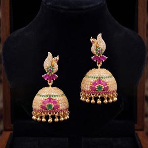 May include: Gold-toned earrings with a floral design. The earrings feature a bell-shaped base adorned with pink and green gemstones, and a decorative top with a peacock-like design. Small gold beads dangle from the bottom.