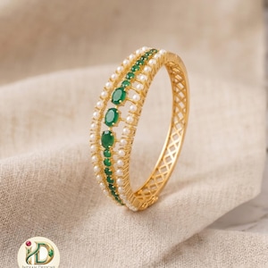 May include: Gold-toned bangle bracelet with a row of oval green gemstones and a row of small white pearls. The bracelet has a textured design on the outer band and is set against a neutral fabric background.