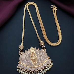 May include: A gold-toned necklace featuring a large, lotus-shaped pendant encrusted with clear stones. The center of the lotus displays a small embossed figure, and the bottom edge is adorned with a row of dangling white beads. The chain is a textured, flexible gold-toned cord.