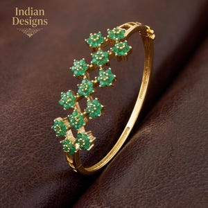May include: A gold-colored bangle bracelet with a floral design. The bracelet features clusters of green gemstones arranged in a floral pattern. The words "Indian Designs" are in the upper left corner.