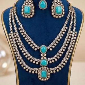 May include: A three-piece jewelry set featuring a necklace, earrings, and a headpiece. The set includes turquoise-colored stones set in a gold-colored metal, with clear, sparkling accents. The jewelry is displayed on a blue velvet stand.